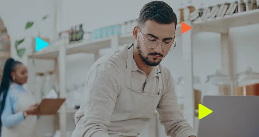 Homem de óculos e avental utilizando um notebook em um estabelecimento comercial, com uma mulher segurando uma prancheta ao fundo desfocado.