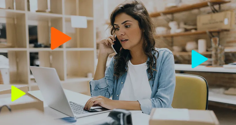 Empreendedora atendendo cliente ao celular e usando notebook em escritório de pequena empresa.