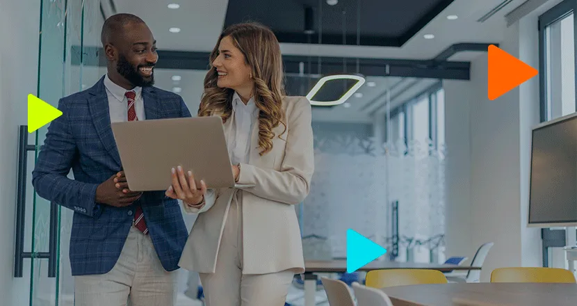 Dupla de executivos sorridentes analisando dados em notebook em escritório moderno com ícones de play coloridos.