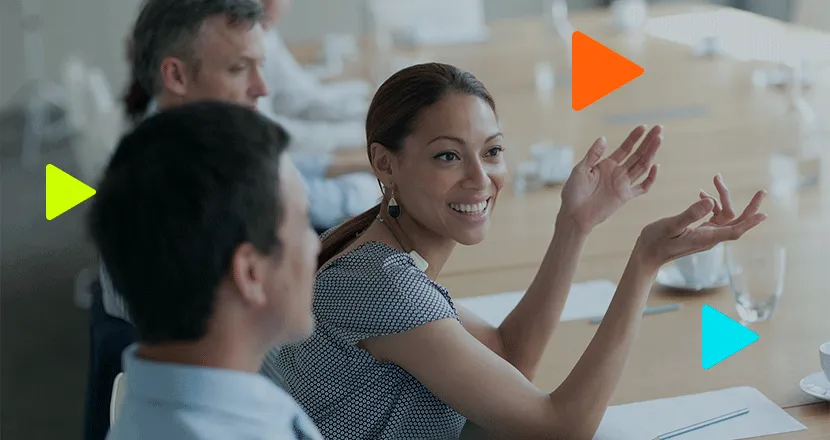 Uma mulher sorridente gesticula durante uma reunião de negócios em uma mesa de conferência com colegas.