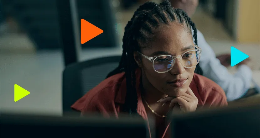 Mulher jovem com tranças e óculos concentrada olhando para monitor, com ícones de play coloridos flutuando ao redor.