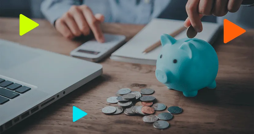 Pessoa colocando moeda em cofrinho de porquinho azul sobre mesa com moedas espalhadas, calculadora e notebook ao fundo.