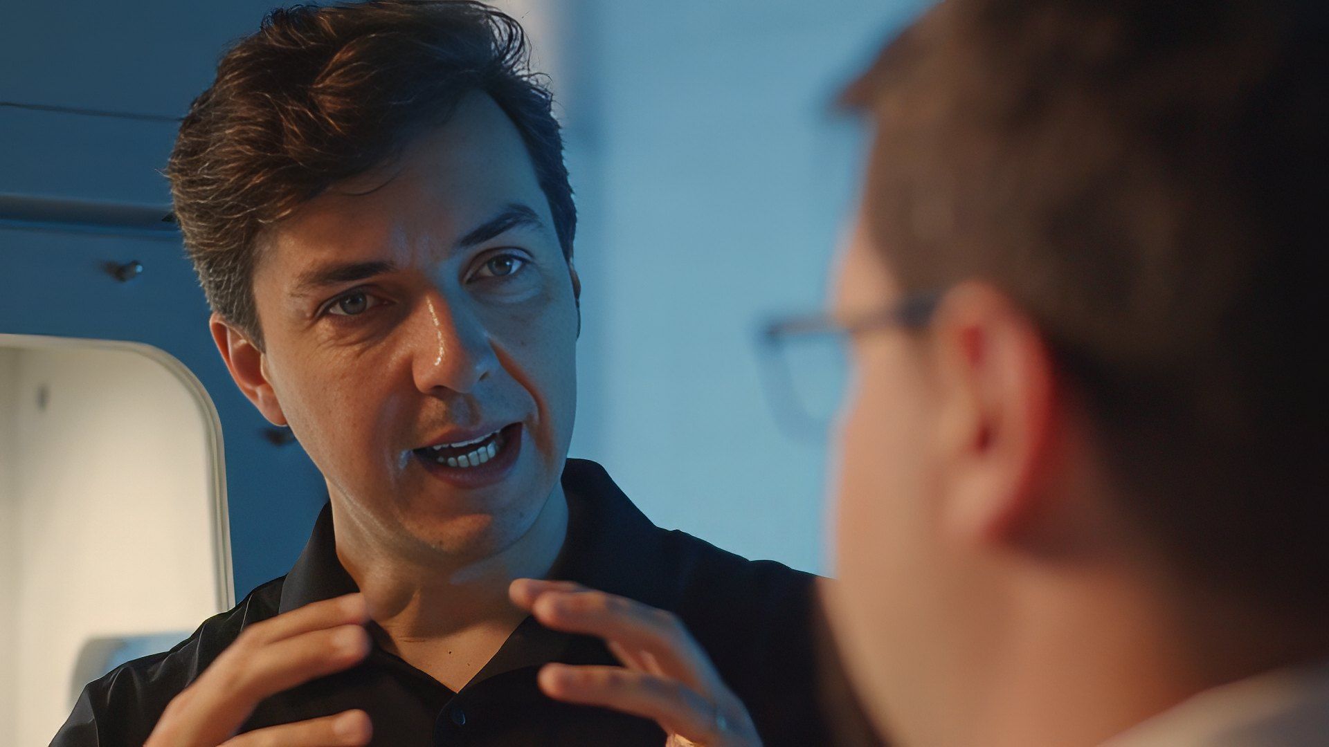 Empreendedor da Hipperquimica gesticula com as maos enquanto explica algo a um colega, em um ambiente industrial com iluminacao azulada ao fundo.